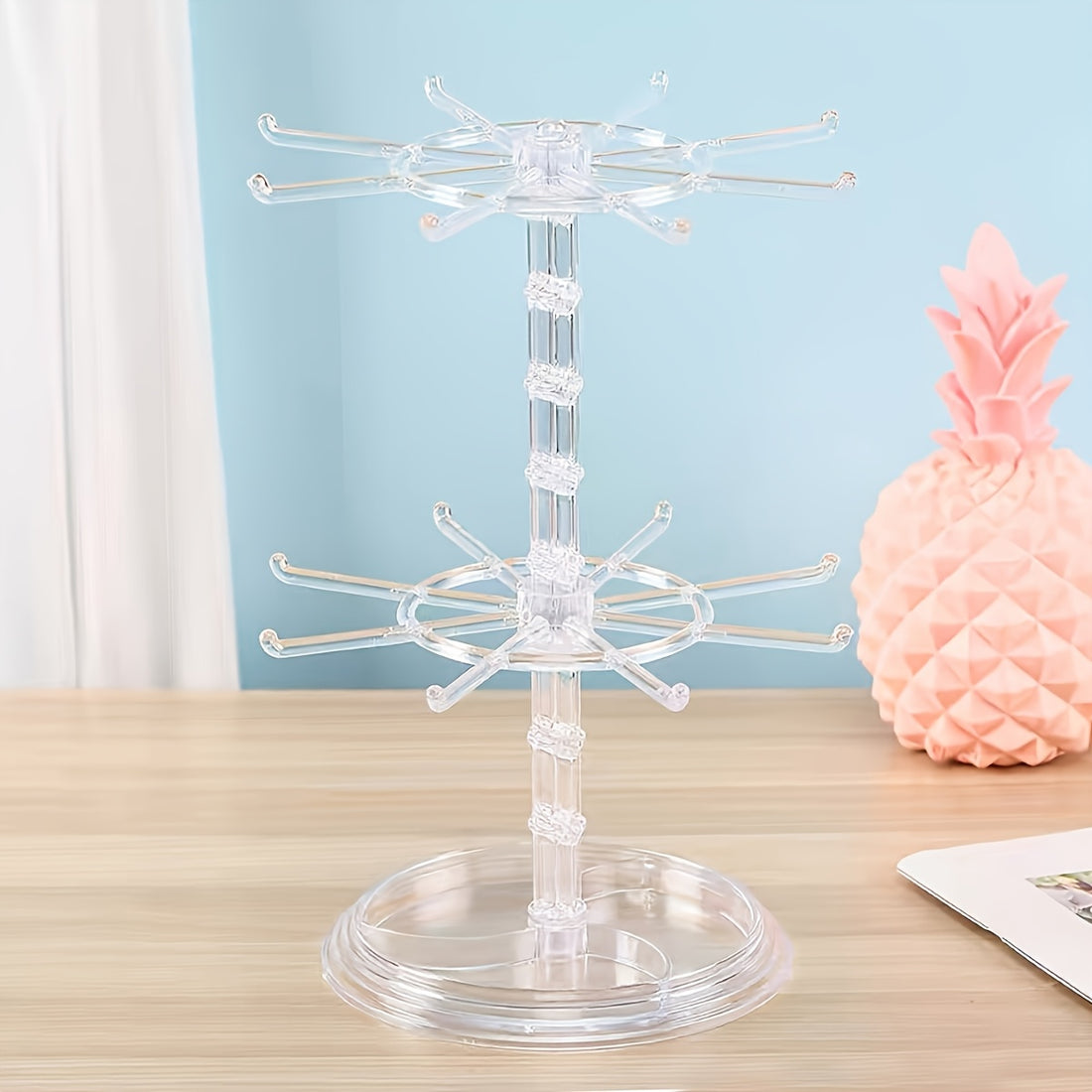 Clear acrylic jewelry display stand on a wooden surface with a pink decorative pineapple in the background.